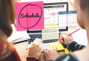 Two people sitting at a laptop with a calendar on its screen. Another calendar is in the photo as well as sticky-notes that have to do lists and a pink note has "schedule" circled on it.