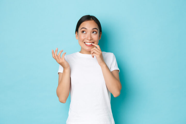 Portrait of dreamy clueless cute asian girl in white t shirt biting fingernail and looking thoughtful up, thinking what to do, making decision, standing blue background and pondering